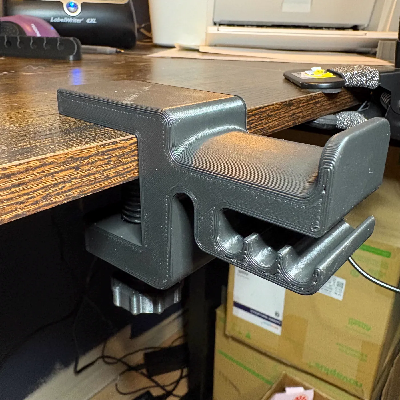 A grey printed headphone holder clamped under the edge of a desk